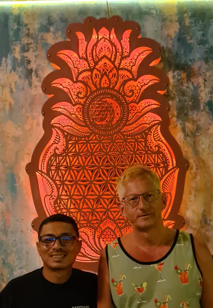 Two men, a dark-haired Thai and a blond European, smile into the camera in front of a red ornamental wall